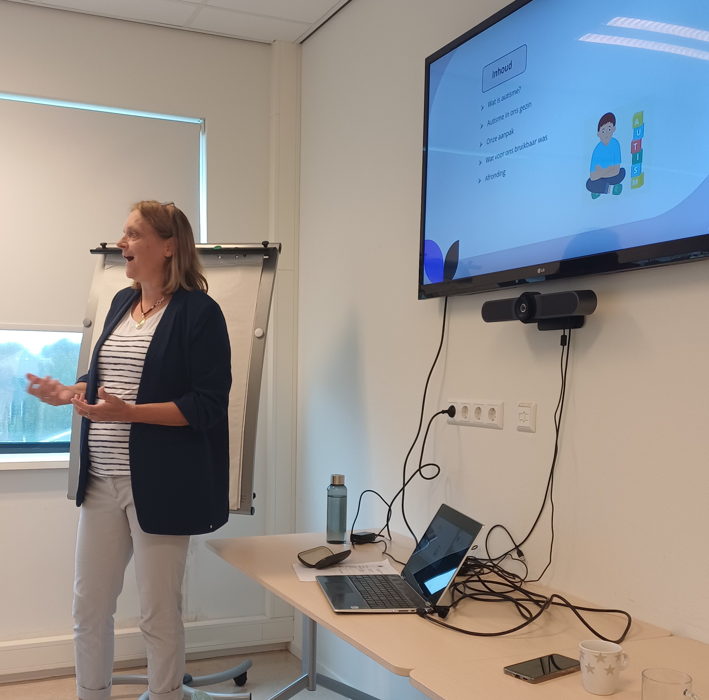 Lotte kraaikamp die een presentatie geeft. Ze staat naar de luisteraars gericht in een open houding. Op de achtergrond een scherm met een dia van een kind met autisme 